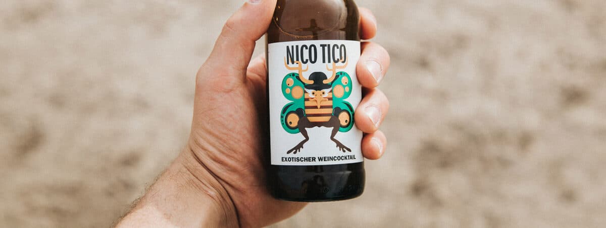Eine braune Flasche der Marke Nico Tico wird in der Hand gehalten. Im Hintergrund ist verschwommen ein leicht bräunlicher Boden zu erkennen.