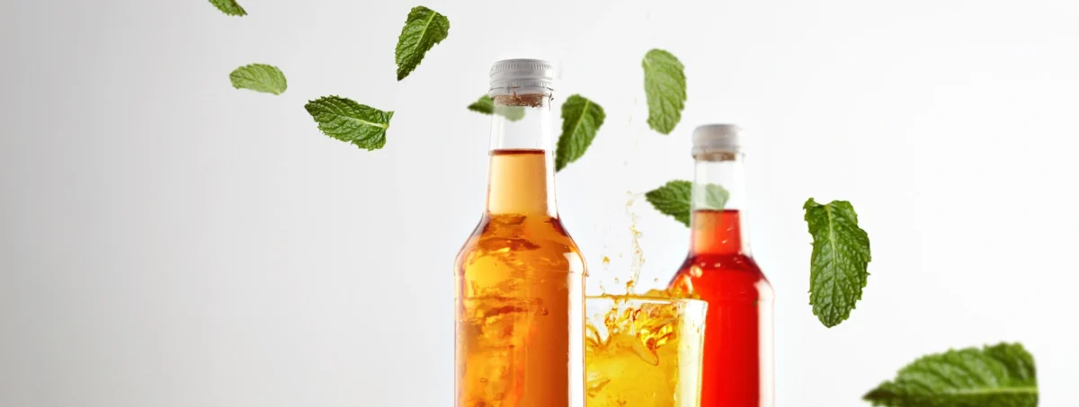 Getränkeentwicklung: Zwei Flaschen von Limonaden, orange und rotfarbig. In der Mitte ein Glas gefüllt mit gelbfarbigem Getränk. Im Hintergrund sind frische und einzelne Minzblätter zu sehen.